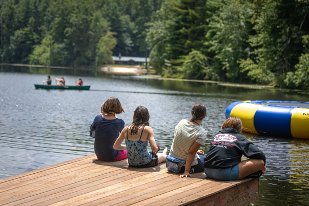 Four students dangling their legs off the edge of a dock at Lake House Academy with a trampoline and students canoeing in the background.