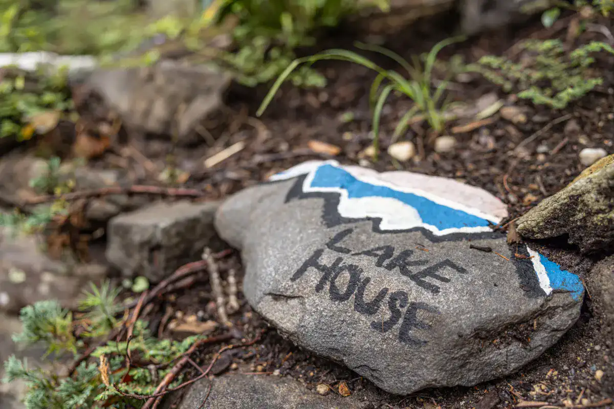 A rock with painted with a blue and white design and the words Lake House sitting in a wooded setting.