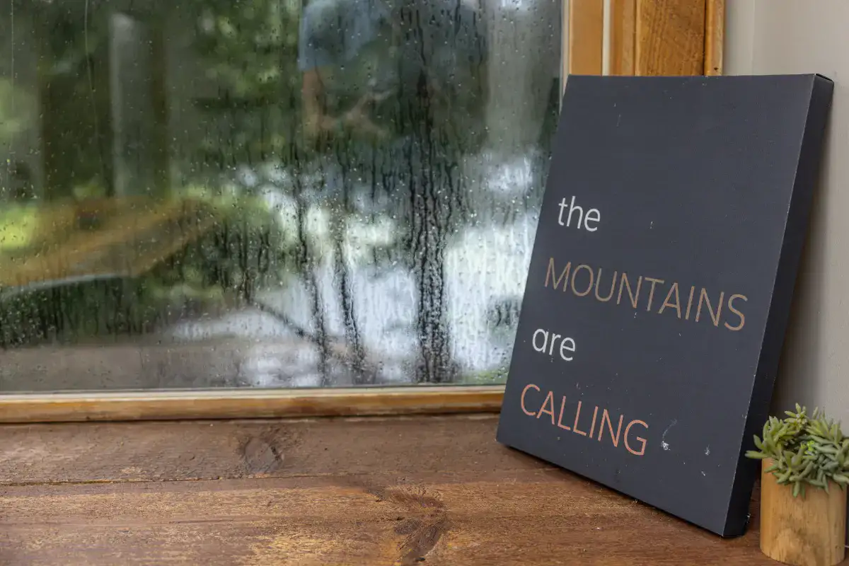 A small sign that reads the Mountains are Calling tucked in a corner against a rainy window.