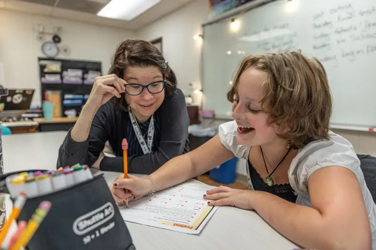 A teacher repositioning her glasses to focus on the work being done by a smiling student at a therapeutic school.