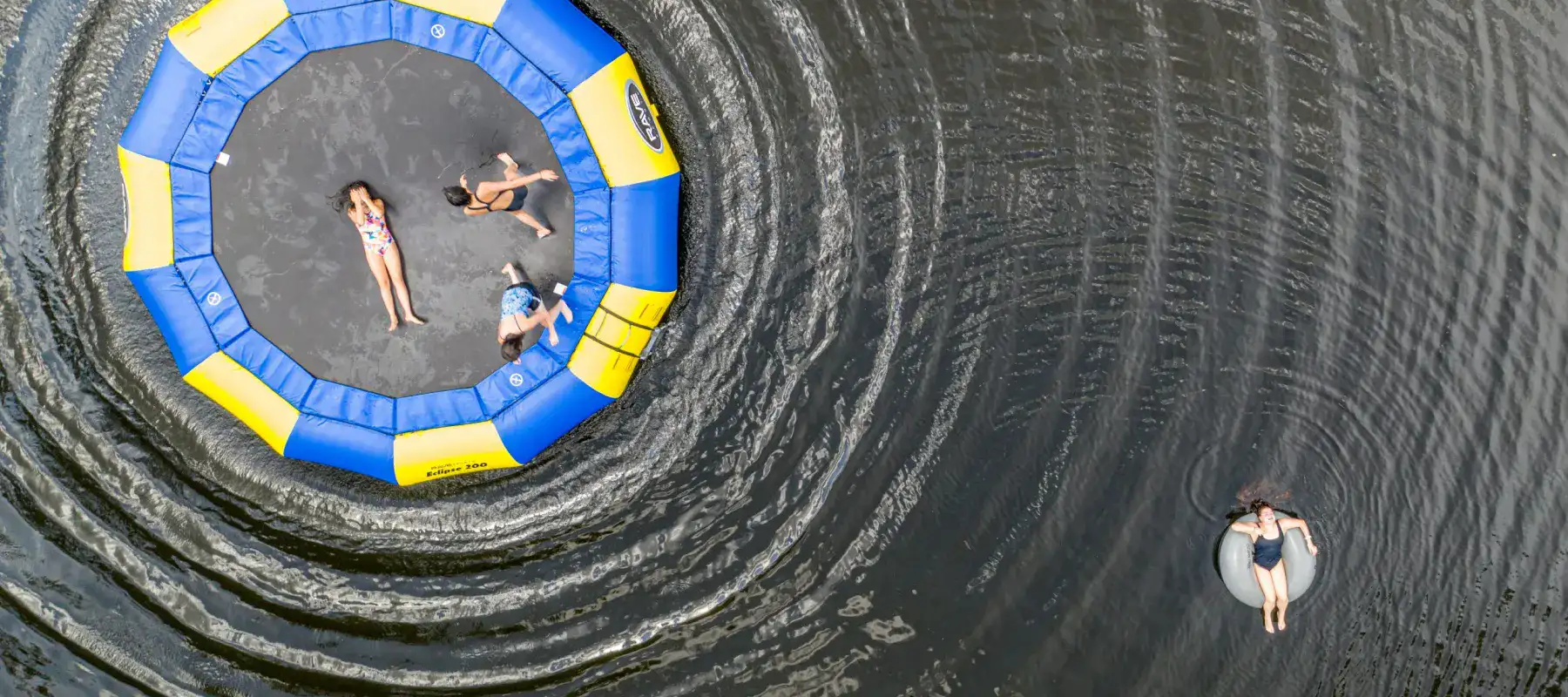 A drone shot of three students playing on a water trampoline and one student floating in a tube on the lake at Lake House Academy.